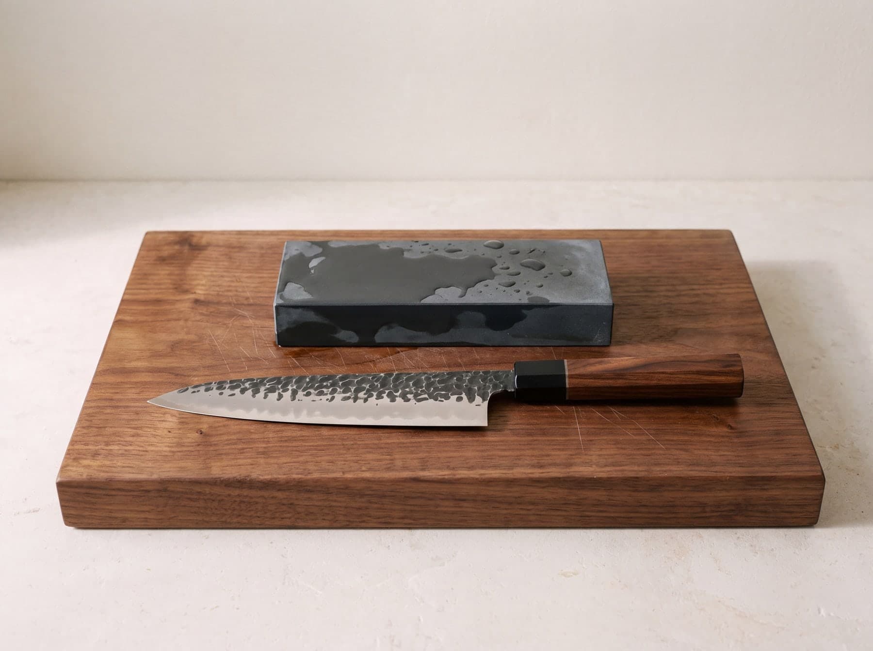 Japanese chef knife with a hammered blade and wood handle next to a wet whetstone on a walnut cutting board